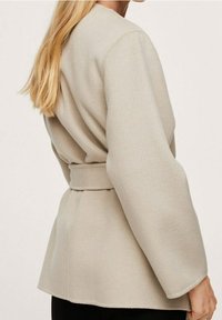 Lichtbeige jas van een zachte stof, voorzien van een wikkelontwerp, brede mouwen en een ceintuur die in de taille is gestrikt. Gladde textuur met minimale details.