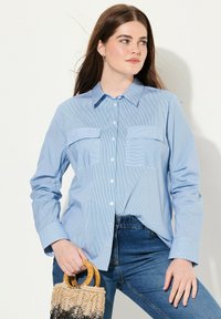 Camicia leggera azzurro chiaro con sottili righe bianche, dotata di due tasche sul petto, maniche lunghe e un colletto classico.
