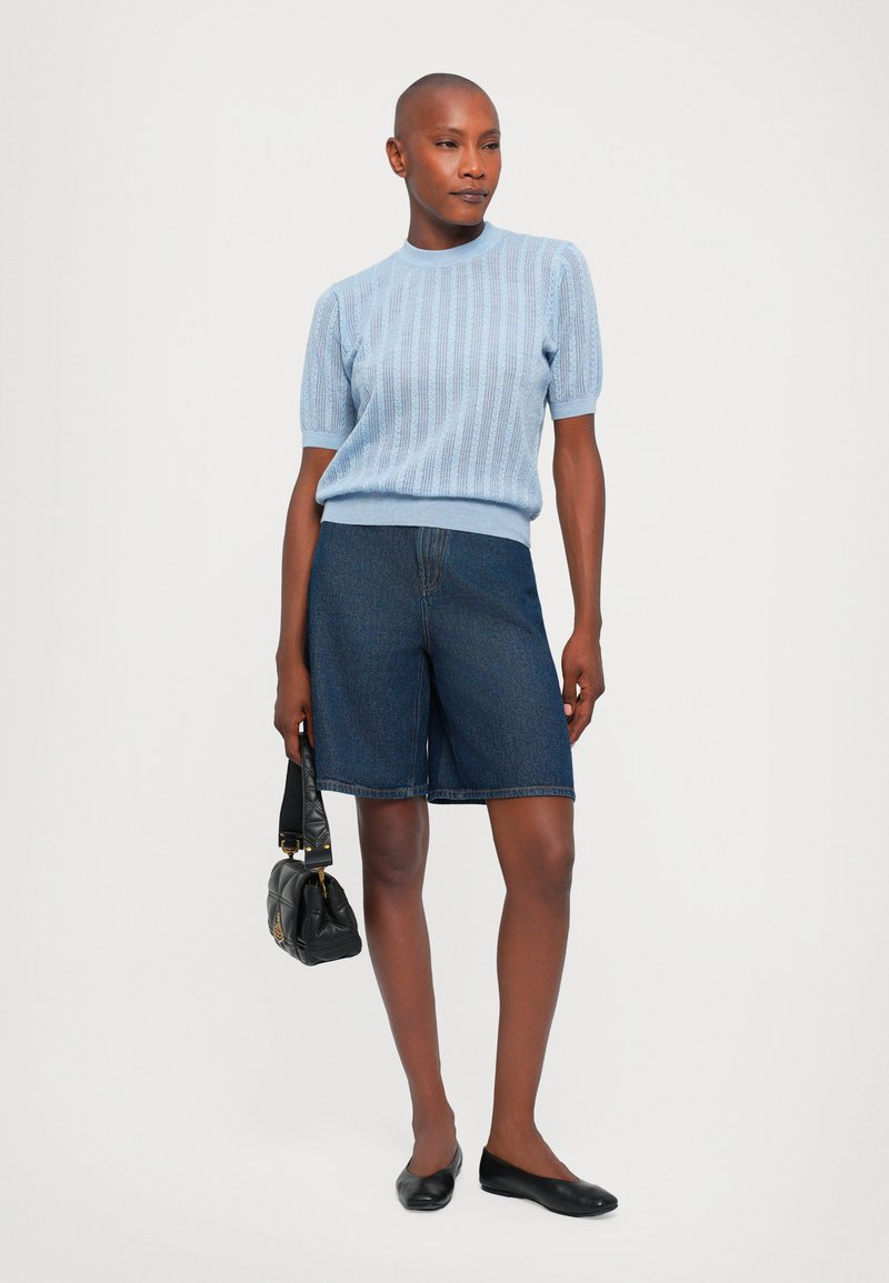 Light blue knitted short-sleeve sweater, dark denim bermuda shorts, black flats, and a small black handbag. Smooth textures and casual design.
