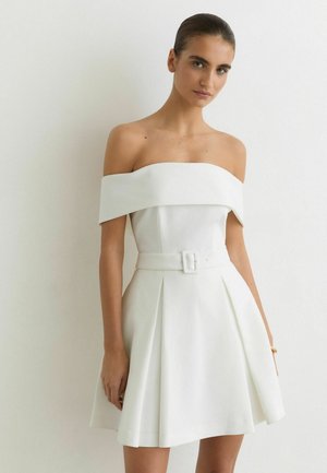 Kvinde iført en hvid off-shoulder kjole med bredt bælte og plisseret nederdel, stående mod en ensfarvet hvid væg.