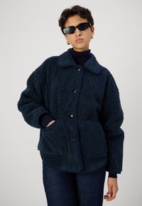 Giacca in pile blu navy con colletto, bottoni frontali e due tasche. Il tessuto ha un aspetto texturizzato e morbido. Indossata sopra un dolcevita.