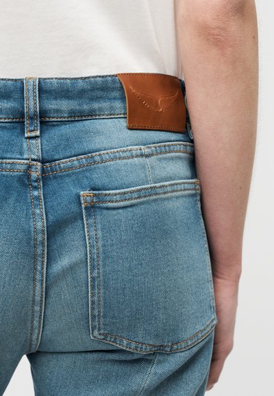 Φωτεινό γαλάζιο τζιν με δερμάτινο πατς στο πίσω μέρος, με καθαρές γραμμές και ραφές σε αντίθεση. Φαίνεται τσέπη στο πίσω μέρος στην δεξιά πλευρά.