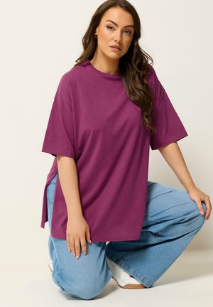 Mujer con cabello largo y castaño arrodillada, vistiendo una camiseta morada holgada, jeans de pierna ancha azul claro y zapatillas blancas, fondo neutro.