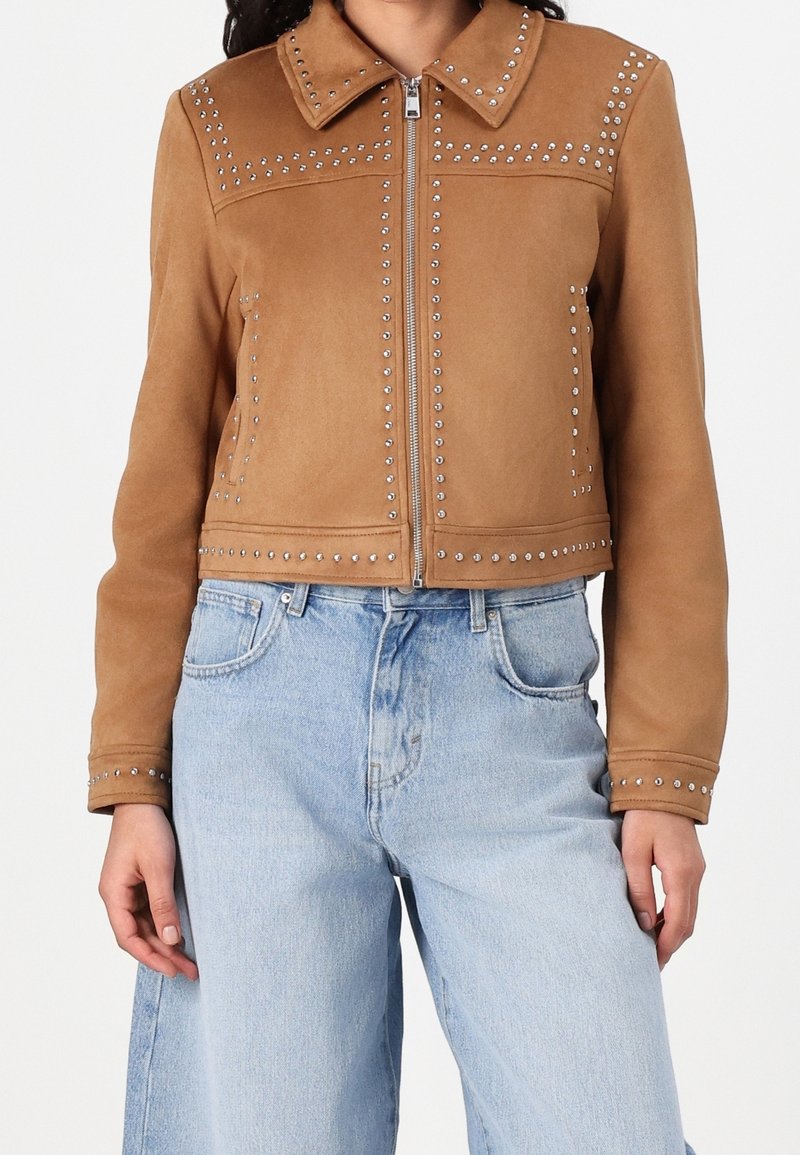 Veste courte en daim marron avec des clous argentés le long des coutures et du col, portée avec un jean bleu clair taille haute.