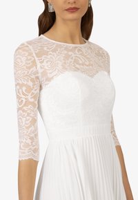 Robe blanche avec un corsage en dentelle à motif floral et des manches trois quarts, complétée par une jupe plissée.