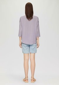 Helles lila, locker sitzendes Oberteil mit drei Viertelärmeln, kombiniert mit hellblauen Denim-Shorts und beigen Riemchensandalen. Einfarbiger Hintergrund.
