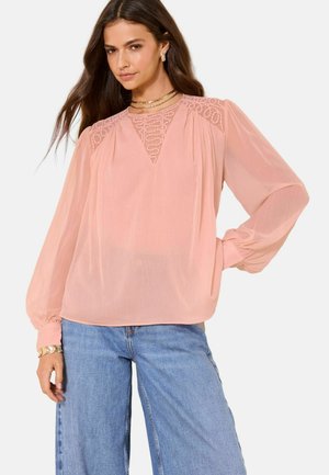 Žena v ružovej priehľadnej blúzke s čipkovými detailmi a naberanými rukávmi, v kombinácii so svetlomodrými džínsami s širokými nohavicami.