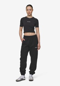 Zwart cropped t-shirt met witte tekst "PEGADOR", gecombineerd met zwarte joggingbroek met elastische boorden en logo. Schoenen zijn grijs met zwarte strepen.
