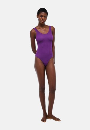 Femme debout pieds nus dans un maillot de bain une pièce violet sans manches et côtelé, sur fond uni clair.