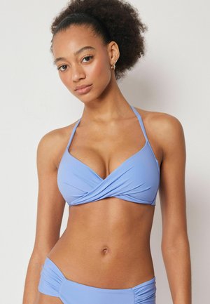 Kvinde med naturligt krøllet hår iført en lyseblå halterbikini, stående foran en ensfarvet hvid baggrund.