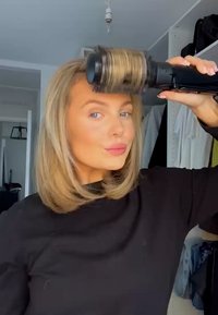 Strumento per capelli nero con un cilindro rotondo ricoperto di setole, utilizzato su capelli biondi chiari di lunghezza spalla. Sullo sfondo ci sono mensole con vestiti.