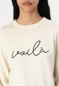 Pull de couleur crème avec une texture tricotée et un col rond, arborant le mot "voilà" en broderie cursive noire sur la poitrine.