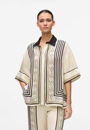 Kortærmet, cremefarvet bluse med sort krave, prydet med lodrette striber og abstrakte mønstre. Knappet foran med sidelommer.