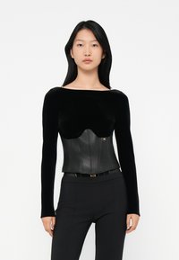 Black long-sleeve top combining soft velvet and structured leather corset. Features a curved bust line with a gold logo accent. Fitted design.
