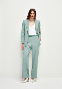 Světle modrý sáčko s pevnou strukturou a kalhoty širokého střihu, doplněné bílým topem a zeleným pásem. Bílé boty na blokovém podpatku.