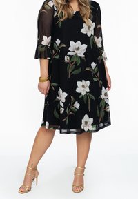 Černé šaty s květinovým vzorem, které zdobí bílé magnolie. Průsvitné tříčtvrteční rukávy a rozšířený lem, doplněné zlatými podpatky.