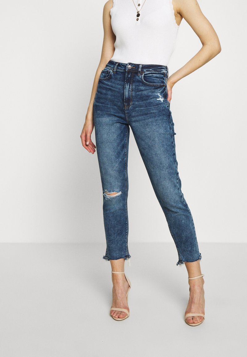 Femme portant un jean bleu déchiré taille haute, un haut blanc sans manches, des colliers superposés et des sandales à talons hauts nude avec des lanières.