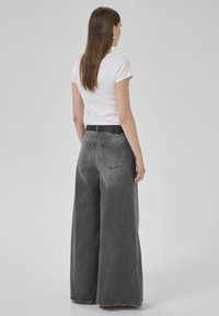 Woman standing facing away wearing a white short-sleeve top and high-waisted wide-leg faded black jeans with a black belt.