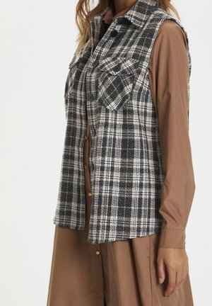 Nainen pukeutunut mustavalkoruudulliseen napitettuun liiviin pitkähihaisen ruskean mekon päällä, sivukuva olkapäästä puolivälikkoon reidestä.