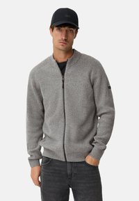 Lysegrå ribstrikket cardigan med lynlås lavet af blødt materiale, med høj krave og lynlås, kombineret med mørke jeans og en kasket.