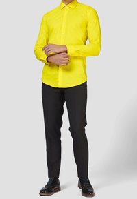 Camisa amarilla de manga larga con cuello clásico, frente abotonado y diseño liso, combinada con pantalones negros y zapatos de vestir.