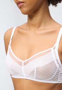 Κοντινό πλάνο ενός ατόμου που φοράει λευκό διχτυωτό σουτιέν με υφασμένα σημεία και ρυθμιζόμενους ιμάντες, εμφανίζοντας το πάνω μέρος του στήθους και την περιοχή του λαιμού.