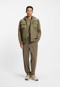 Oliven grøn puffer vest med to forlommer, parret med en matchende oliven langærmet skjorte og beige bukser, afsluttet med lyse sneakers.