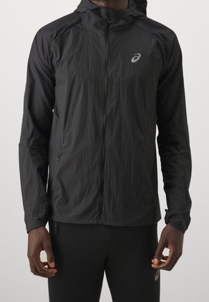 Homme portant une veste légère noire à capuche zippée avec un petit logo réfléchissant de la marque sur la poitrine et un pantalon noir assorti.
