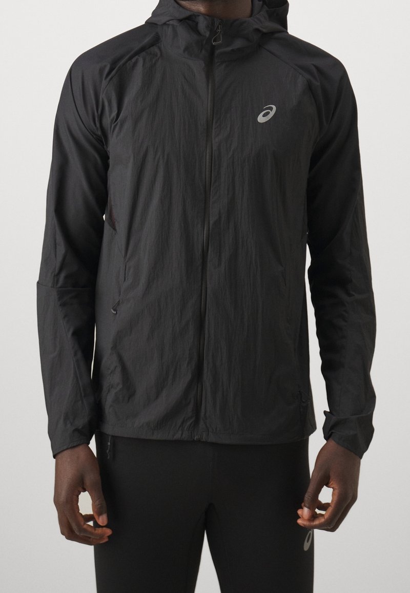 Homme portant une veste légère noire à capuche zippée avec un petit logo réfléchissant de la marque sur la poitrine et un pantalon noir assorti.