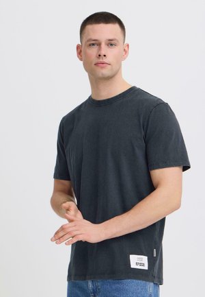 Mladý muž v černém tričku s krátkým rukávem a modrých džínách, stojící se sepnutýma rukama, neutrální výraz, jednobarevné světlé pozadí.