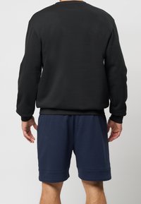 Hombre vistiendo un sudadera negra de manga larga y pantalones cortos hasta la rodilla en azul marino, de pie de espaldas con los brazos ligeramente doblados.