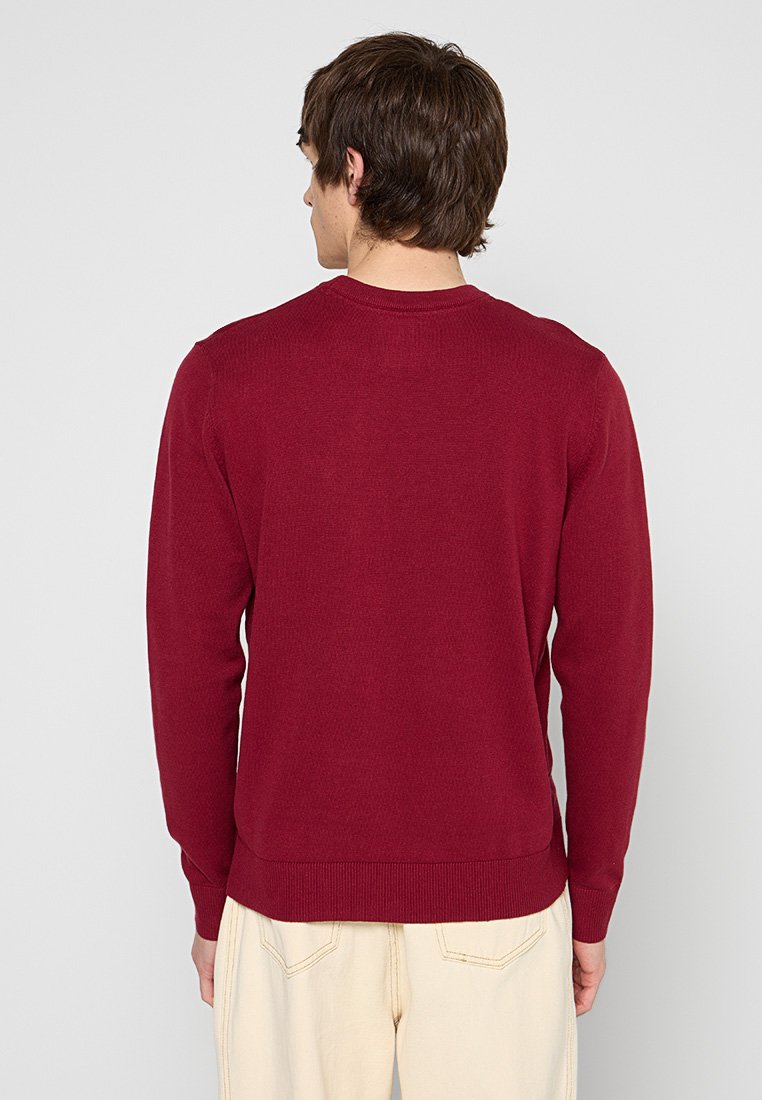 Pull en tricot bordeaux avec un col rond, des manches à poignets côtelés et une coupe ajustée. Le tissu présente une texture uniforme, idéale pour des tenues superposées.