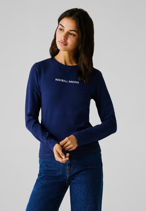Ženska z dolgimi temnimi lasmi, oblečena v modro dolgo majico z napisom "RED BULL RACING" in modre kavbojke, stoji z rokami prekrižanimi.