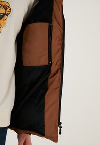 Sort faux fur vest med en brun stofforing, der har en lynlåslomme. Vestens tekstur er glat, og den har et kontrasterende farveskema.