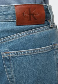 Blugi din denim albastru deschis cu o pată de piele maro pe talie, având logo-ul "Calvin Klein" embosat și inițialele "CK".