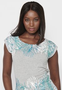Camisa gris de manga corta con patrones florales en teal y blanco en el cuello, con un corte relajado y una textura de tela suave.