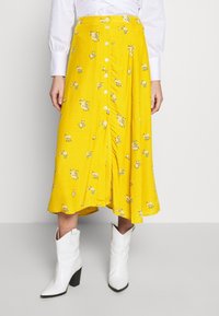 Jupe midi fleurie jaune avec un devant à boutons, ornée de boutons blancs et d'une texture douce. Portée avec des bottes de cowboy blanches.