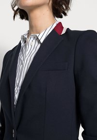 Granatowa marynarka z podkreślonymi ramionami, zapięciem na jeden guzik z przodu oraz kieszenią na piersi. Wewnętrzny kołnierz ozdobiony jest akcentem w kolorze bordowym i białym. Tkanina o fakturze.