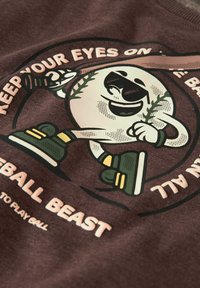 Tessuto marrone con un personaggio dei cartoni animati, una palla da baseball con braccia e gambe, testo slogan attorno in bianco e verde, elementi di design giocosi.