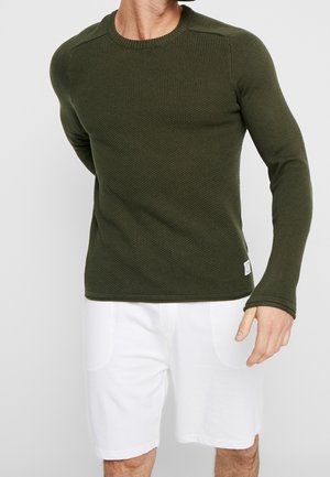 Homme portant un pull en maille texturée vert olive et un short blanc, debout avec un bras derrière le dos sur un fond uni.