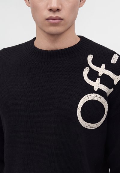 Jeune personne portant un pull en tricot noir avec de grandes lettres blanches cursives brodées sur la partie supérieure gauche de la poitrine.
