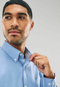 Šviesiai mėlynas marškinis su mygtukais, kurio vidinė apykaklė turi langelį. Medžiaga atrodo lygi, su mygtukais apykaklėje ir rankogaliuose.