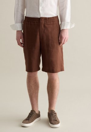 Shorts en lin marron avec une taille élastique, longueur genoux, présentant une finition texturée. Portés avec des baskets en daim marron.