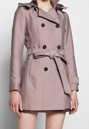 Femme portant un trench-coat mauve à double boutonnage avec des boutons noirs et une ceinture nouée à la taille, par-dessus une jupe courte ou une robe.