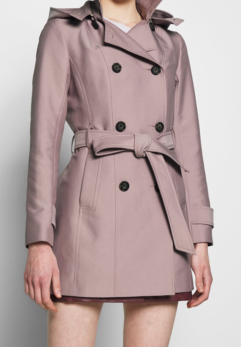 Femme portant un trench-coat mauve à double boutonnage avec des boutons noirs et une ceinture nouée à la taille, par-dessus une jupe courte ou une robe.
