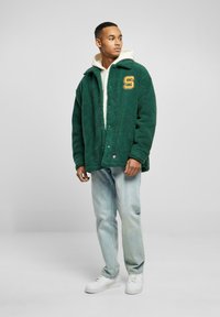 Grønn fleecejakke med krage, frontknapper, gult bokstav "S"-lap, båret over en hvit hettegenser og lyseblå jeans. Hvite sneakers.