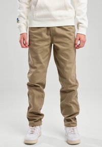 Beige Baumwollhosen mit lockerer Passform, mit Seitentaschen und umgeschlagenen Säumen, kombiniert mit weißen Sneakers und einem hellen Hoodie.