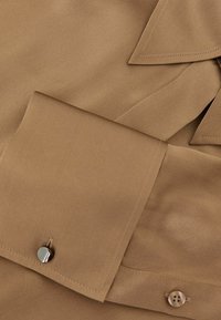 Ruda šilkinė palaidinė su lygiu paviršiumi, su apykakle, mygtukų papuošimais ir tvarkingai siuvinėtomis kraštinėmis. Dėmesys skiriamas blizgiems mygtukų detalėms.