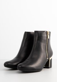 Botines de cuero negro hasta el tobillo con punta redonda, cremallera lateral y acentos en metal dorado. Presentan un tacón cuadrado con una base dorada en contraste.