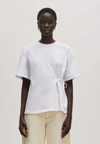 Witte katoenen t-shirt met korte mouwen, voorzien van een gerimpelde zijdetail vastgemaakt met een trekkoord. Het ontwerp is ontspannen en iets oversized.